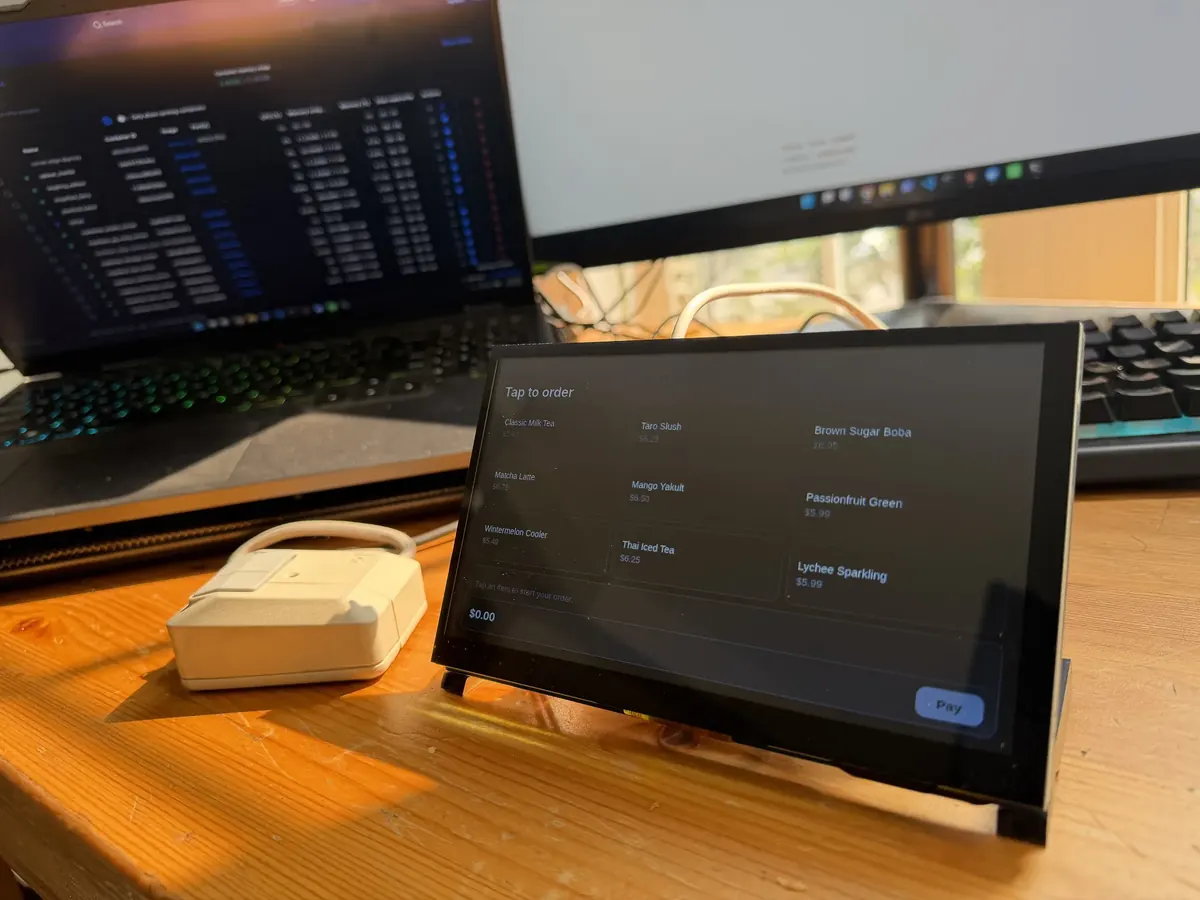 The Corner bench kiosk, a small touchscreen on a wooden bench, showing a tap-to-order drink menu