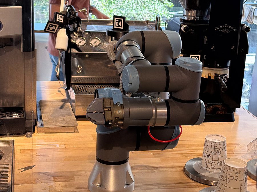 A grey robot arm behind a glass screen on a butcher-block counter, an espresso machine at left and a grinder at right, paper cups at the counter’s edge