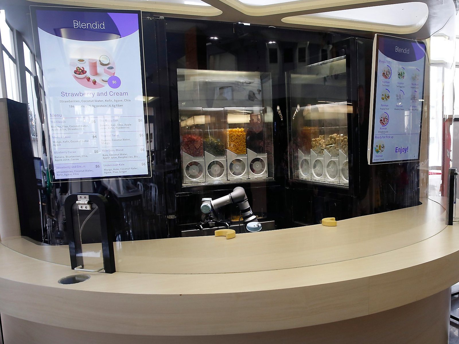 The Blendid kiosk: a curved cream counter under a glass canopy, menu screens either side, ingredient dispensers and a robot arm behind glass