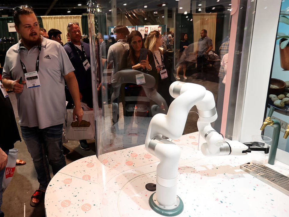 Trade-show visitors with lanyards watch a white robot arm inside Bobacino’s curved glass bar, gold taps at right