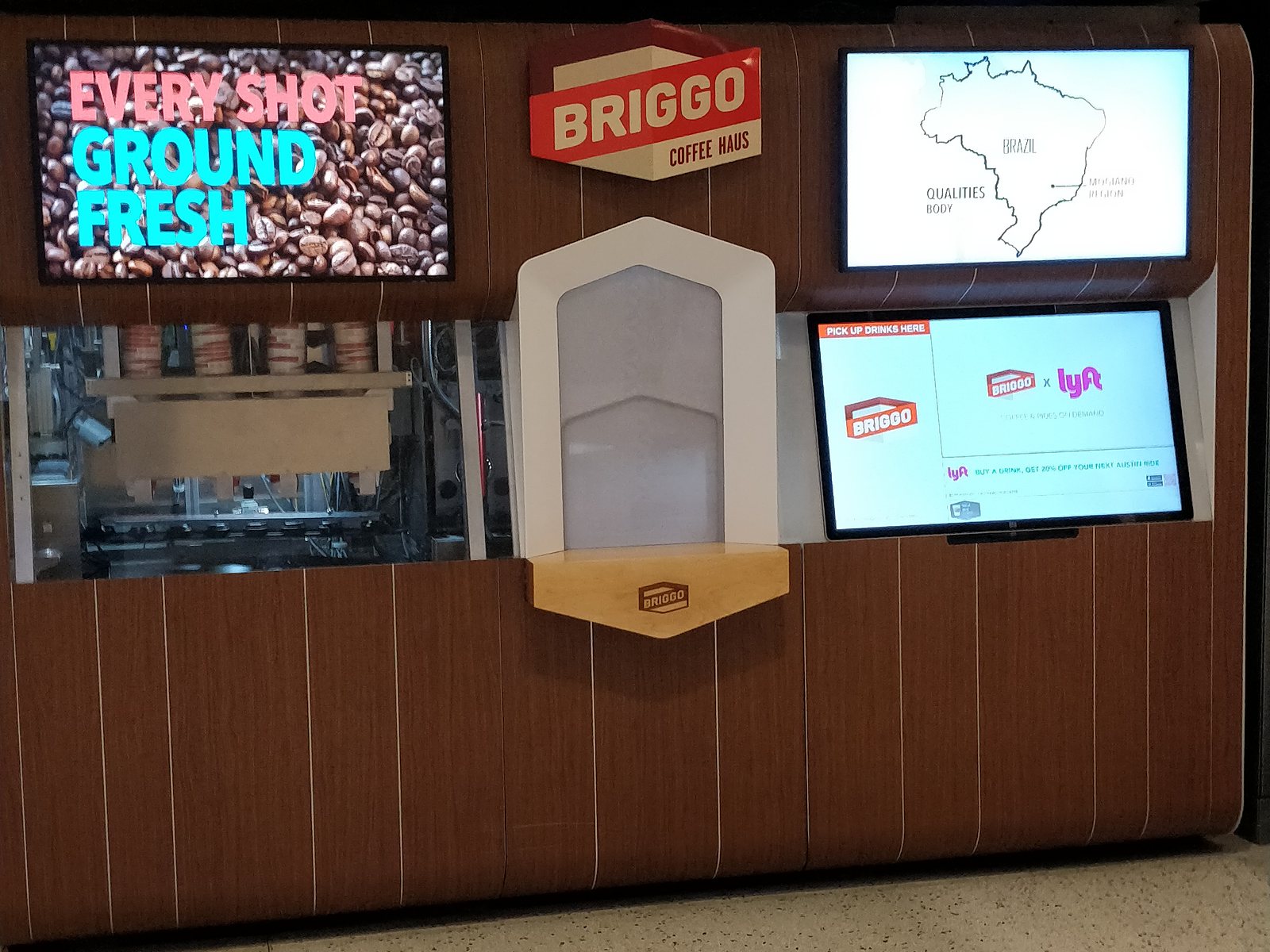A wood-slatted Briggo Coffee Haus kiosk set into an airport wall, a window onto the machine at left, a pickup cubby in the middle and order screens