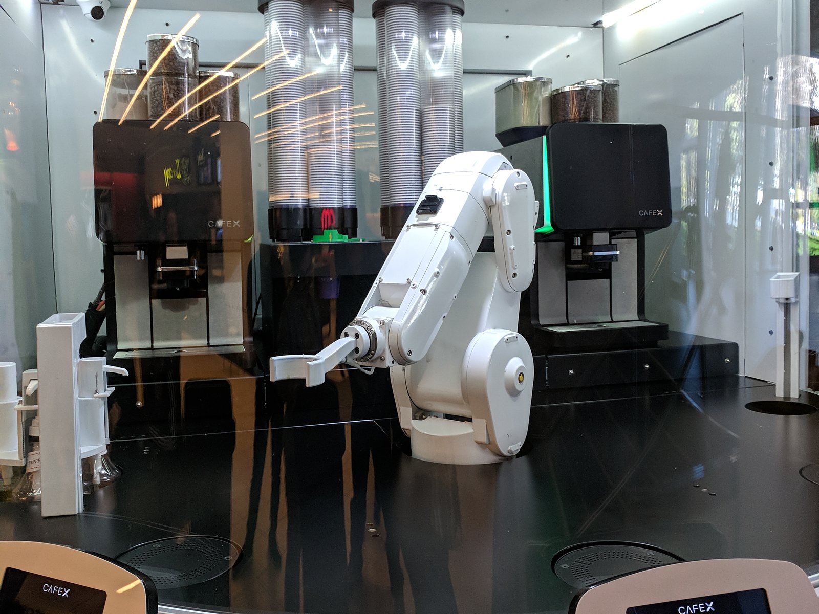 A white industrial robot arm between two espresso machines and two towers of cups, behind curved glass