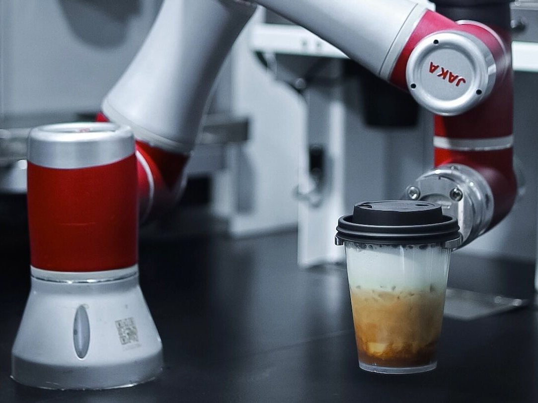 A red and white robot arm setting a lidded iced latte down on a dark counter