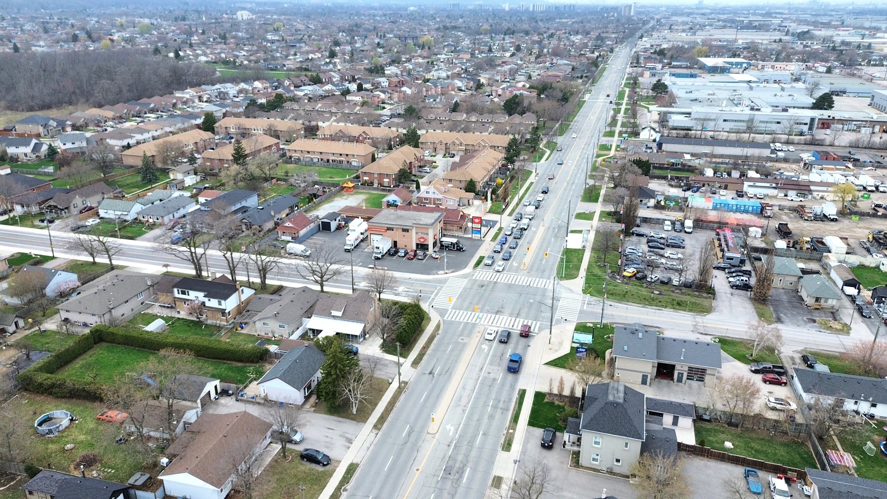 High aerial of the Barton Street East and Dewitt Road junction, the corner building and its lot at centre, townhouse blocks behind and an industrial strip along Barton to the right