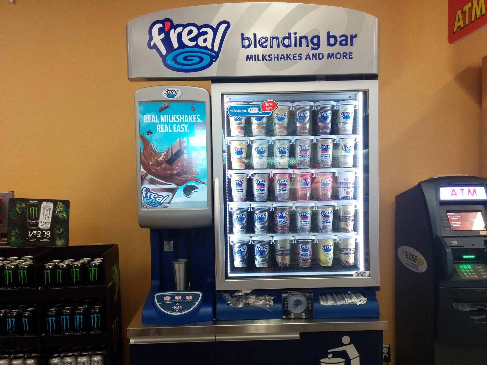 An f’real blending bar in a store: a blender unit with a screen at left and a lit glass-door freezer of f’real cups at right