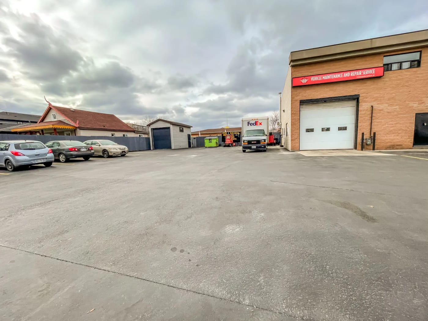 The small rear structure with its roll-up door beyond a row of parked cars, the service building’s east face and bay door at right