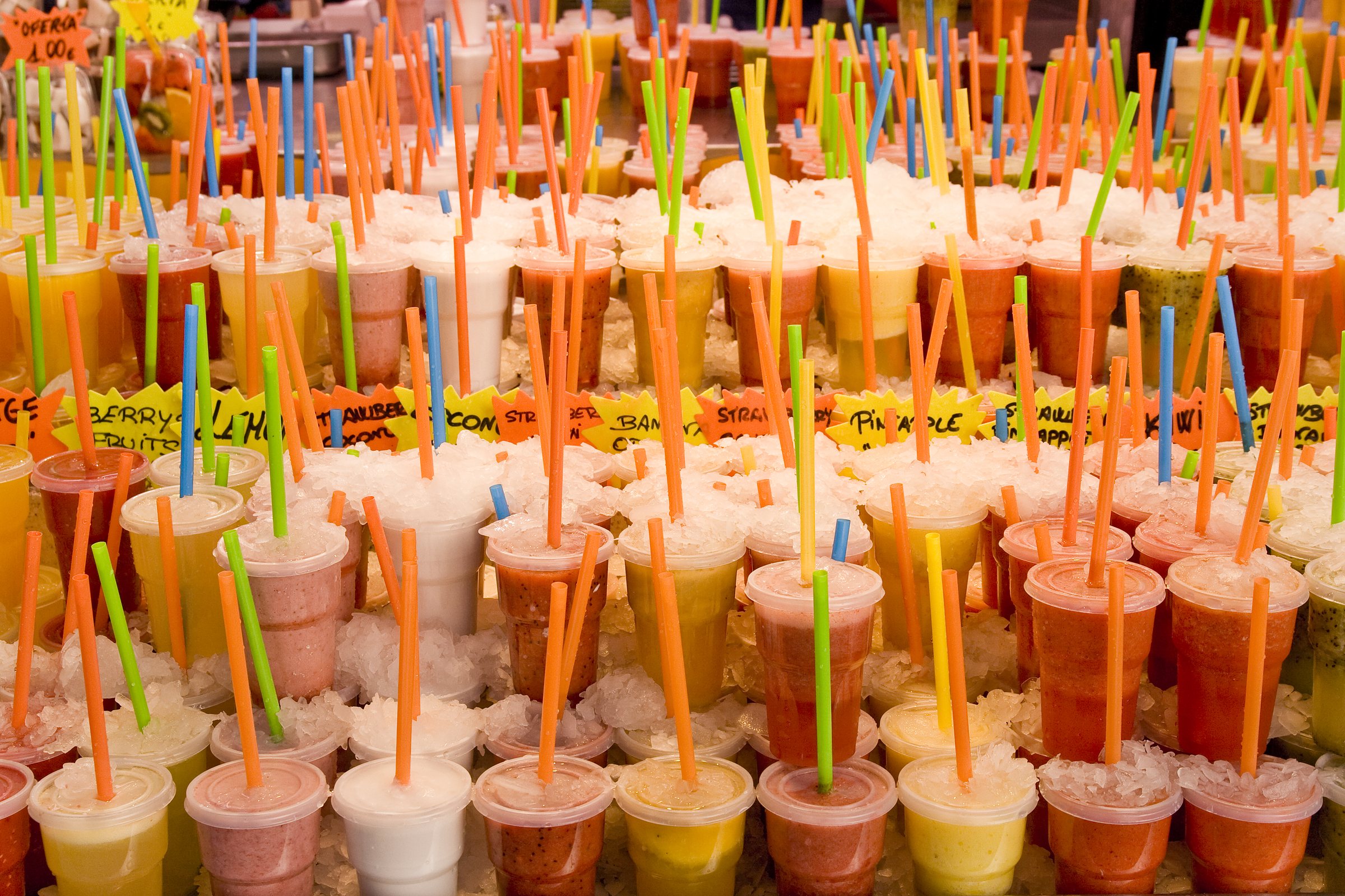 Rows of fruit juices and smoothies in plastic cups with coloured straws, packed in crushed ice at a market stall, with handwritten flavour signs