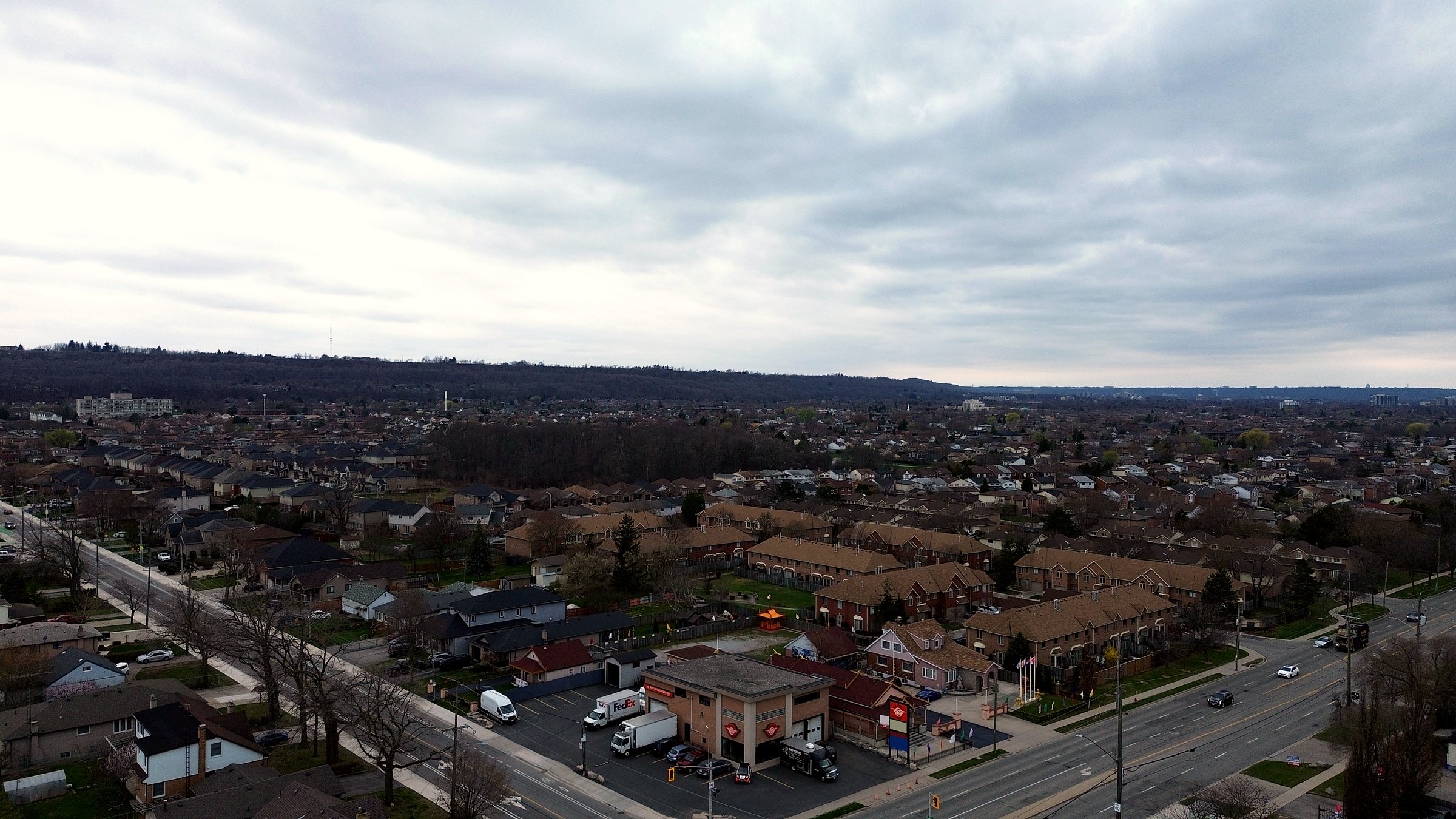 A wide aerial view of the corner and the surrounding neighbourhood under an overcast sky, the escarpment on the horizon