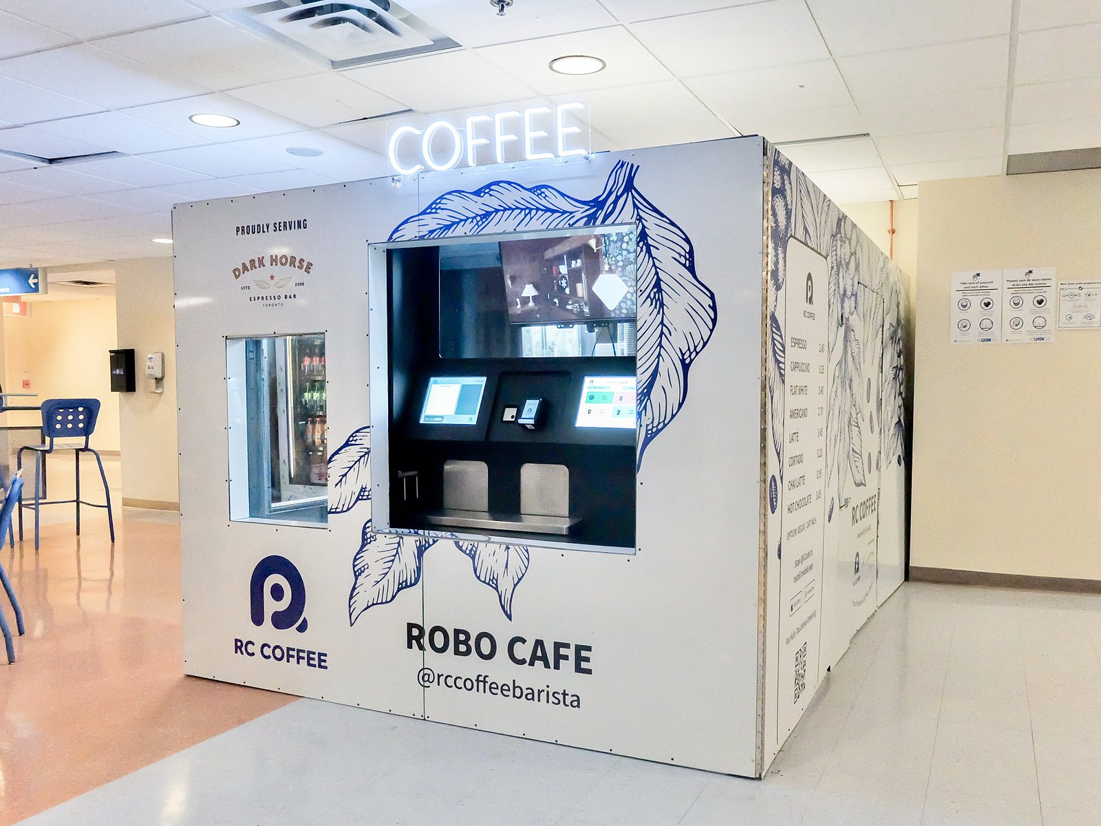 A free-standing white RC Coffee Robo Cafe kiosk with blue leaf drawings and a neon coffee sign, order screens in its window, in a hospital food court