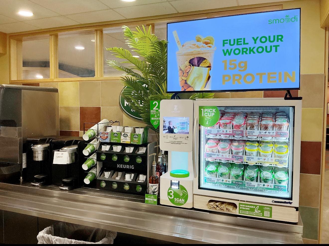 A Smoodi self-serve blending kiosk on a stainless counter, a glass-front freezer of fruit cups beside it and a screen above