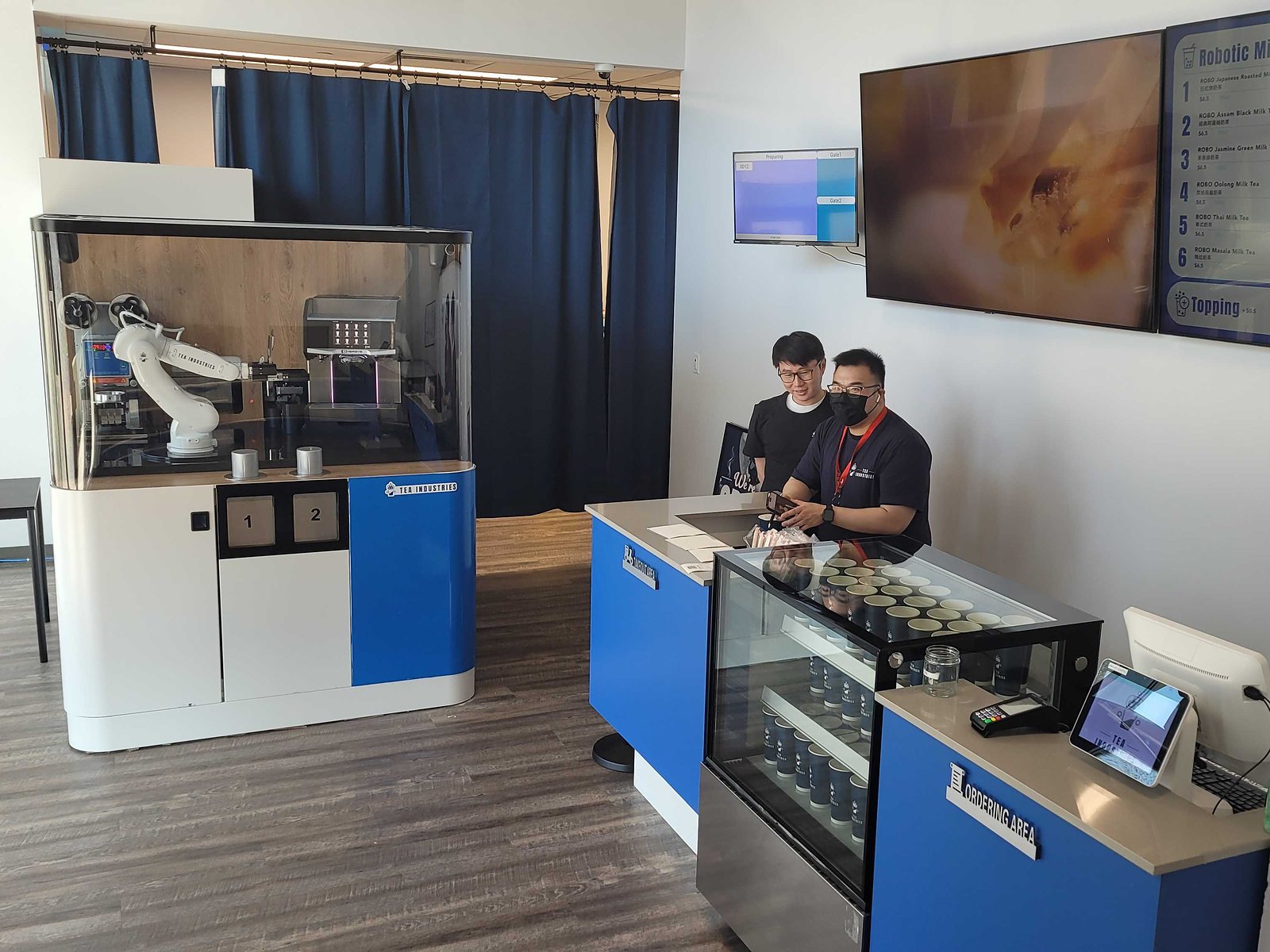 Inside Tea Industries: a white robot arm in a glass cabinet with two numbered pickup doors at left, a blue ordering counter and two staff at right