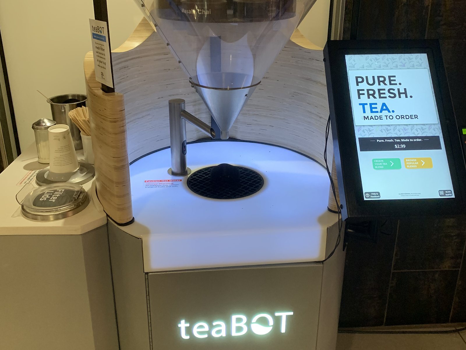 A teaBOT kiosk: a clear cone and funnel over a hot-water spout and a lit cup platform, an order screen at right, the lit teaBOT logo below