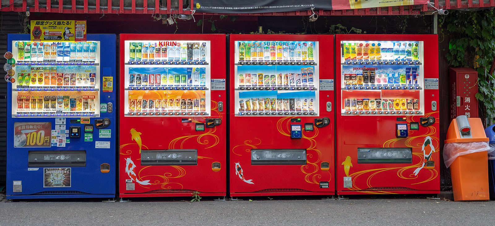 A row of four brightly lit beverage vending machines along a wall in a Tokyo park