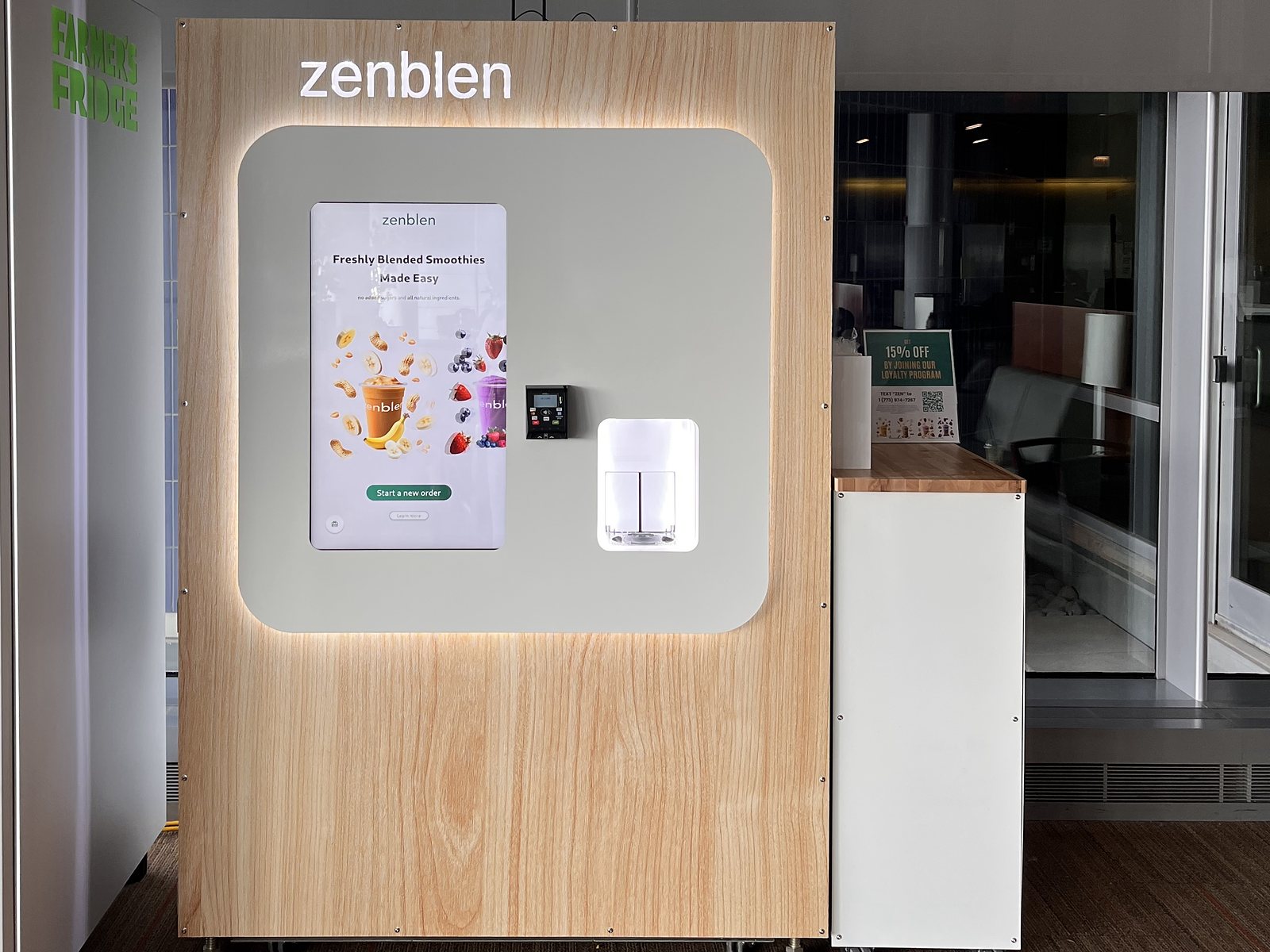 A wood-grain Zenblen kiosk with a lit sign, a touchscreen, a card reader and a lit pickup window, a white side table beside it